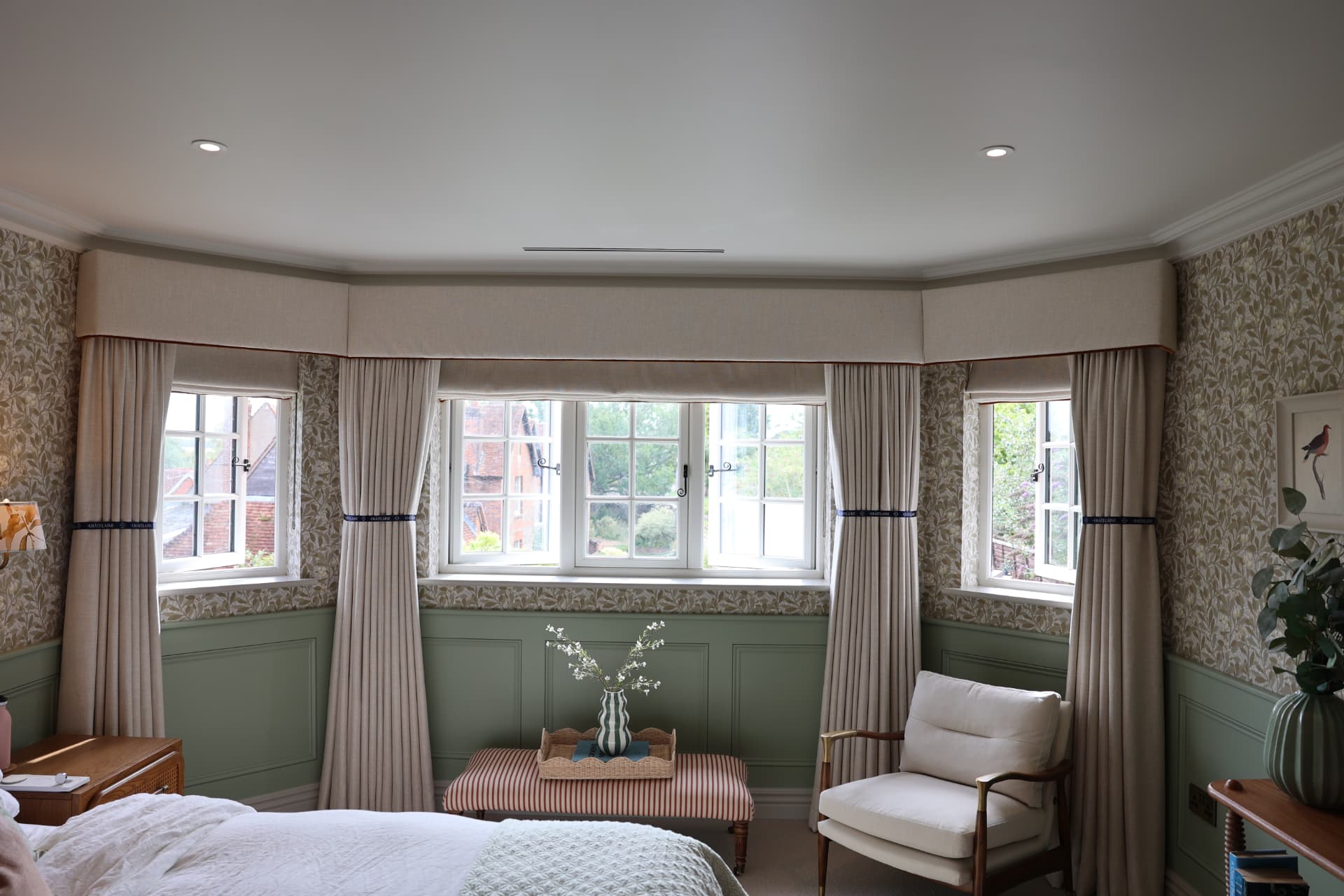 A bay window dressed with a plain oatmeal upholstered pelmet with piped trim, gathered curtains on branded ribbon tie-backs and matching roman blinds, against botanical wallpaper and sage paneling