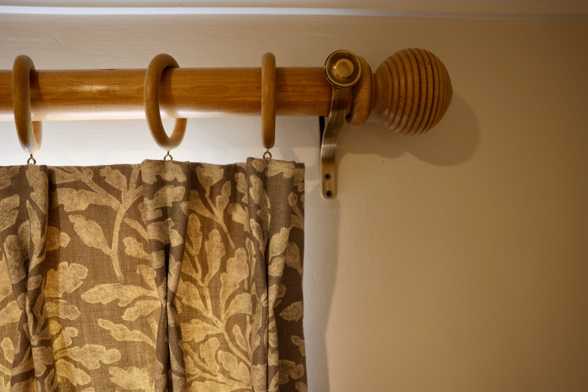 A honey-toned wooden curtain pole with matching rings and a grooved spherical finial on an antique brass bracket, above the head of a leaf-patterned pinch-pleated curtain