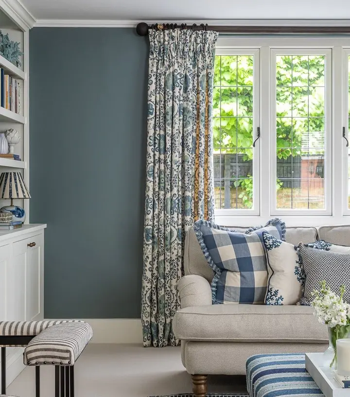 A window corner dressed in full-length pinch-pleated curtains, with a pale sofa, patterned cushions and a striped ottoman